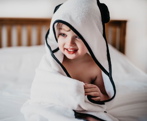 Smiling child sitting on a bed wearing the hooded bamboo towel.